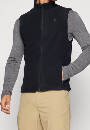 Mann trägt eine schwarze Fleeceweste mit Reißverschluss über einem grauen Langarmshirt und beigen Hosen, mit einer Hand in der Westentasche.