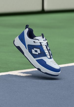 Fehér és kék sportcipő fűzővel, egy teniszpálya közelében, a határvonal felett lebegve.