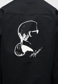 Hvit minimalistisk sideprofil av en kvinne med hestehale og solbriller, med en kursiv signatur under, på et svart plagg.