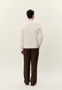 Άνδρας που στέκεται με την πλάτη γυρισμένη, φορώντας λευκό μακρυμάνικο πουκάμισο και σκούρο καφέ παντελόνι, μπροστά σε απλό ανοιχτό φόντο.