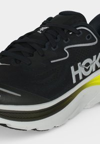 Μαύρο και λευκό παπούτσι τρεξίματος Hoka με μαύρα κορδόνια, επάνω μέρος από δίχτυ και λεπτομέρεια λευκής και κίτρινης σόλας.