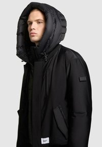 Czarna puchowa kurtka z dużym, ocieplanym kapturem, wyposażona w kieszeń na zamek i naszywkę z logo. Gładka tekstura i nowoczesny design.