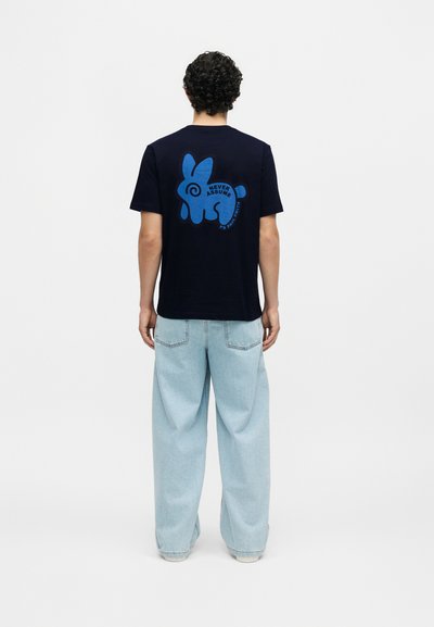 Άνδρας με σγουρά μαλλιά που φοράει μαύρο T-shirt με μπλε αφηρημένο κουνέλι και το κείμενο "Never Assume" στην πλάτη, σε συνδυασμό με άνετο ανοιχτό μπλε τζιν.