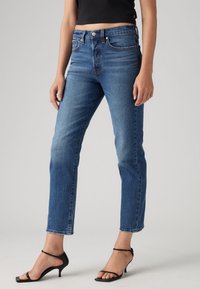 Vaqueros de mezclilla azul con corte recto, con cierre de cremallera y botón, cinco bolsillos y costuras en contraste. Llevados con sandalias de tacón negras.
