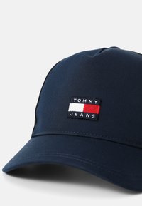 Jūras zilas krāsas cepure no austo auduma. Aprīkota ar taisnstūrveida izšūtu uzlīmi ar "TOMMY JEANS" un sarkaniem, baltajiem un zilajiem akcentiem.