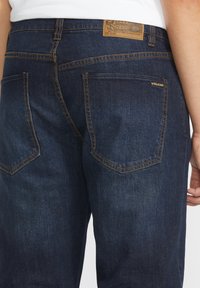 Vaqueros de mezclilla azul oscuro con costuras naranjas en contraste, dos bolsillos traseros y una etiqueta de marca de cuero en la cintura.
