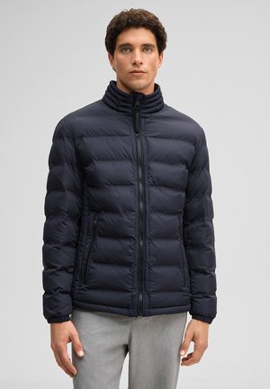 Mężczyzna ubrany w ciemnogranatową, pikowaną kurtkę zapinaną na zamek z wysokim kołnierzem oraz jasnoszare spodnie, stojący na tle jednolitego, jasnego tła.