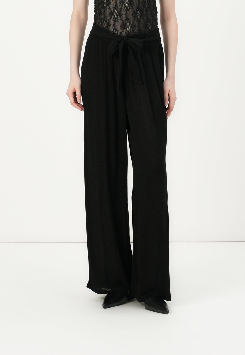 Pantalones anchos negros con una textura suave, con una cintura atada y un diseño fluido. Combinados con una blusa de encaje negra.