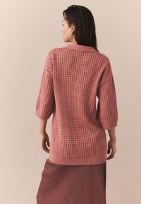 Rožinės spalvos megztinis su laisvu stiliumi, trijų ketvirčių rankovėmis ir tekstūruotu atviru pynimo raštu. Pabaigos detalės – ribinė juosta apačioje ir rankogaliai.