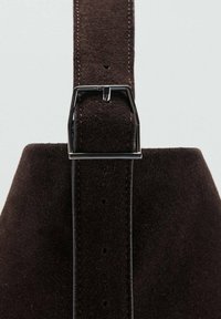 Correa de ante marrón con una hebilla de metal plateada, con costuras y agujeros visibles para el ajuste. La superficie texturada es suave y rica en color.