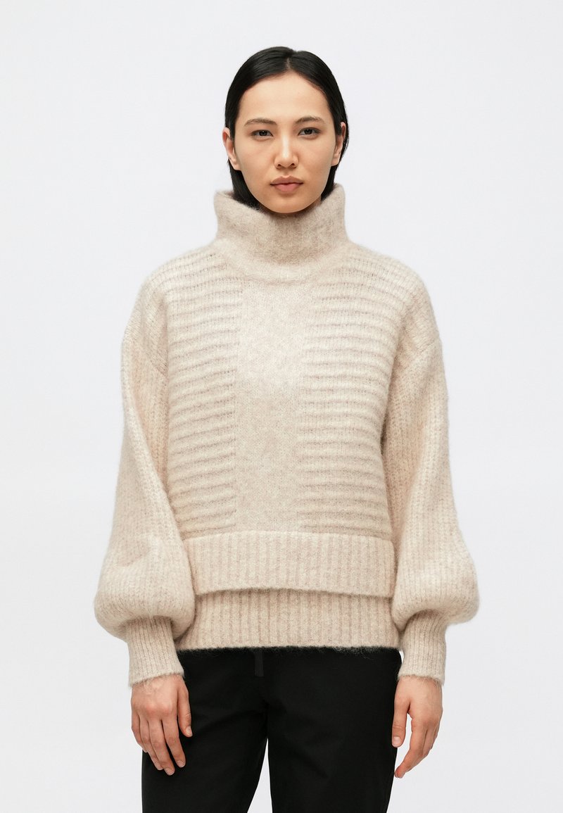 Beige rullekrave sweater med ribbet tekstur og oversized ærmer, der har en bred kant og blødt, fluffy materiale.