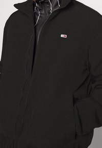 Black zip-up jacket featuring a high collar, front pockets, and a small logo patch. Made of lightweight material with a smooth texture.