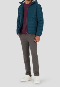 Homme portant une veste matelassée teal, une chemise bordeaux, un pantalon gris et des baskets blanches se tenant contre un fond léger uni.