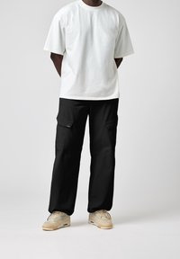 Weißes Baumwoll-T-Shirt mit kurzen Ärmeln, kombiniert mit schwarzen Cargo-Hosen mit seitlichen Taschen. Cremefarbene Sneakers runden das Outfit ab.