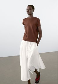 Brun kortærmet t-shirt, hvide vide bukser og sorte flip-flops med røde accenter. Glat stof, afslappet silhuet, minimalistisk design.