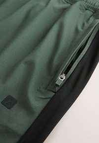 Green and black pants with an elastic waistband and a zippered side pocket showing the zipper pull and fabric texture.