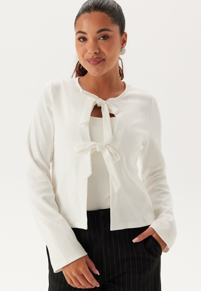 Cardigan bianco a costine con scollo rotondo e dettagli a fiocco davanti. Maniche lunghe e design aderente. Indossato sopra una maglietta chiara.