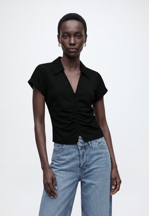 Kvinde iført en sort bluse med korte ærmer og rynket front samt lyseblå højttaljede jeans, står foran en ensfarvet grå baggrund.