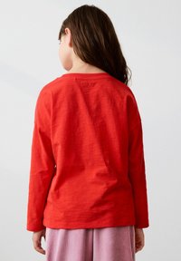 Enfant avec de longs cheveux bruns, portant une chemise à manches longues rouge et un pantalon léger violet, se tenant dos à un fond uni.