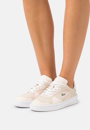 Paire de baskets basses beiges avec des lacets et une semelle blanches, arborant un logo crocodile vert sur le côté, portées sur des jambes nues.