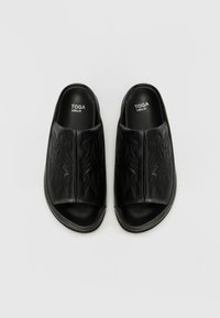Pantofi negri slip-on cu un model floral embosat, textură de piele netedă, design cu vârf deschis și talpă rezistentă. Eticheta de brand "TOGA VIRILIS."