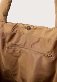 Hnědá látková taška s vnitřním prostorem ukazujícím zipovou kapsu a uzávěr na patentku pod širokými ramenními popruhy.