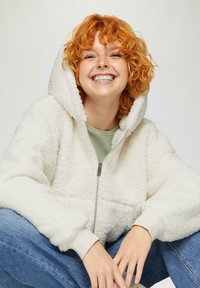 Weißer, flauschiger Reißverschluss-Hoodie mit großer Kapuze und gerippten Bündchen. Trägt man über einem grünen Oberteil, dazu blaue Jeans.