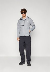 Lys grå isolert jakke med svarte detaljer, frontglidelås og justerbar hette, kombinert med mørke bukser og svarte joggesko.