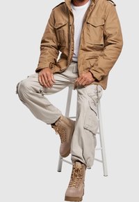 Khaki bunda se čtyřmi předními kapsami, béžové cargo kalhoty a hnědé šněrovací boty. Hladká tkanina s podrobným šitím a zpevněnými švy.