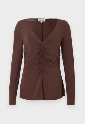 Femeie cu păr lung, purtând un top maro cu mâneci lungi cu pliuri, pantaloni bej cu picioare largi și manșete în carouri, pantofi negri și o geantă neagră.