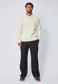 Pull léger crème avec col, cinq boutons et bords côtelés, associé à un pantalon large noir et des chaussures foncées.