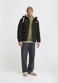 Veste doudoune noire avec capuche, doublure en fausse fourrure beige contrastante, patch de logo beige, associée à un pull olive et à un jean large gris.