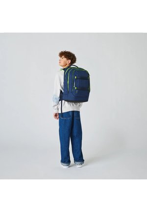 Junge trägt weißen Pullover und blaue Jeans, trägt einen großen dunkelblauen Rucksack mit grünen Akzenten, steht vor einem schlichten hellen Hintergrund.