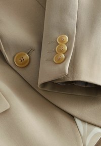 Chaqueta beige ajustada con cuatro botones grandes y redondeados de color claro en la manga. Tejido suave y texturizado con un ligero brillo.