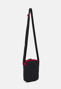 Borsa a tracolla nera, forma rettangolare, realizzata in tessuto. Presenta una parte superiore con zip rossa e una tracolla regolabile. Texture liscia, design minimale.