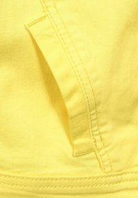 Gelbes Gewebe mit glatter Textur und sichtbaren Stichen. Verfügt über eine kleine Tasche und eine Naht am unteren Rand.