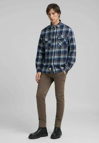 Camisa de franela a cuadros en color marino, azul y marrón, con dos bolsillos en el pecho, cierre de botones y mangas largas. Combinada con pantalones marrones y botas negras.