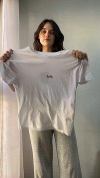 Femme tenant un t-shirt blanc avec « lala BERLIN » imprimé en petites lettres noires sur la poitrine, debout à l’intérieur près d’un rideau transparent.