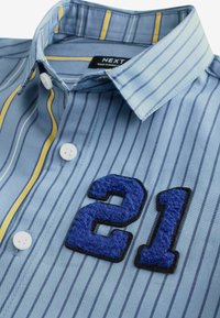 Chemise bleue à rayures boutonnée avec col, présentant des rayures verticales jaunes et bleu foncé ainsi qu'un patch bleu texturé avec le numéro 21 sur la poitrine.