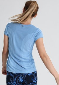 Camiseta deportiva azul claro de manga corta, tejido texturizado, paneles de malla y un pequeño detalle de logo en la parte trasera. Combinada con leggings estampados.