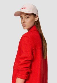 Rote Wolljacke mit hochgekrempelten Ärmeln; hellrosa Baseballmütze mit rotem Text "OLIVER." Das Model posiert seitlich und zeigt die Designdetails.