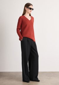 Sweter w kolorze rdzy z prążkowanym wzorem i dekoltem w kształcie v, zestawiony z czarnymi, wysokim talią spodniami w stylu szerokich nogawek oraz czarnymi butami na slip-on. Modelka nosi czarne okulary przeciwsłoneczne.