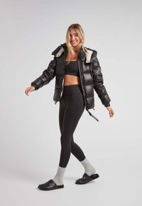 Giacca a vento nera con cappuccio foderato in pelliccia bianca, chiusura con zip e orlo elasticizzato; abbinata a leggings neri e ciabatte nere con calzini grigi.