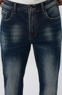 Jean en denim bleu foncé délavé avec bouton frontal et poches, porté avec un haut blanc visible à la taille.