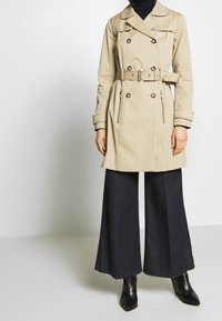 Trench coat beige de doble botonadura con cinturón, con un amplio collar, bolsillos laterales y costuras detalladas, combinado con pantalones oscuros de pierna ancha.