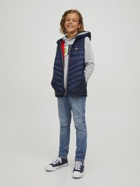 Námornícka vypuklá vesta s kapucňou cez šedú mikinu, spárovaná s modrými džínmi a námorníckymi teniskami. Obsahuje zips a bočné vrecká.