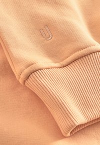 Felpa color pesca in tessuto con polsini a costine. Presenta un discreto logo ricamato a forma di "u". Superficie texturizzata con materiale liscio e elastico.