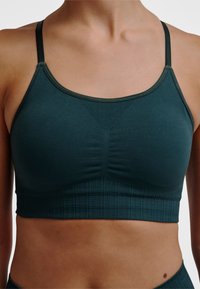 Donkergroene sportbeha met dunne bandjes, gestructureerde stof en een geraffineerd detail in het midden. Geribbelde band aan de onderkant biedt ondersteuning.