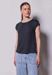 Top negro de manga corta con escote redondo y un detalle fruncido en el frente. Tejido suave con un corte holgado, combinado con jeans de color azul claro.