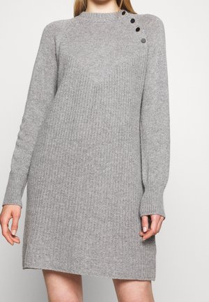 Vrouw draagt een lange grijze gebreide sweaterjurk met lange mouwen en vier zwarte knopen langs één schouder.